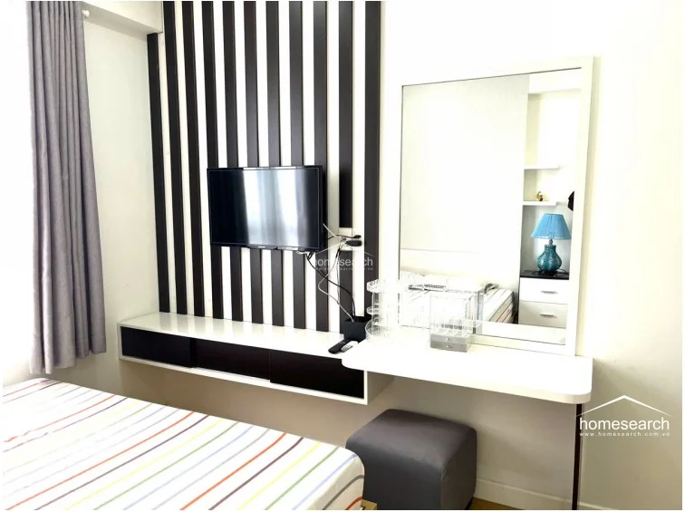This bedroom features a modern, compact layout with a built-in vanity and wall-mounted TV console featuring a bold black-and-white striped accent wall. The room is finished with light-colored walls and a matching grey stool, offering a clean, contemporary aesthetic.