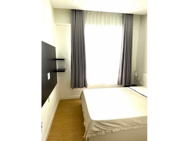 This angle shows the bedroom's flow, emphasizing the wood-look flooring and the wall-mounted shelving unit opposite the bed. The space feels bright and airy, with the large window and neutral walls creating a comfortable, functional sleeping environment.