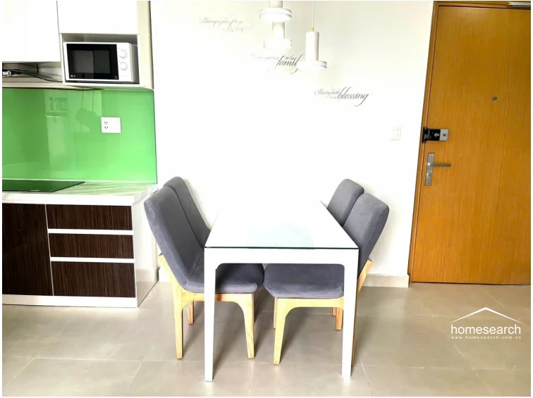 This dining area is situated adjacent to the kitchen, which features dark wood cabinetry and a vibrant green glass backsplash. A modern white dining table with four grey upholstered chairs sits under contemporary pendant lighting, near the main entrance door. The overall condition is well-maintained, utilizing a bright and functional color scheme that complements the open floor plan.