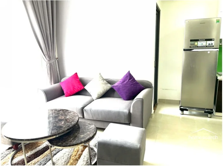 This angle captures the transition from the living space to the kitchen area, featuring a large, stainless steel Panasonic refrigerator. The living room remains consistent with its grey sofa and marble-topped coffee table, set against a backdrop of light tile flooring. The open-plan layout allows for a seamless flow between the relaxation zone and the kitchen.