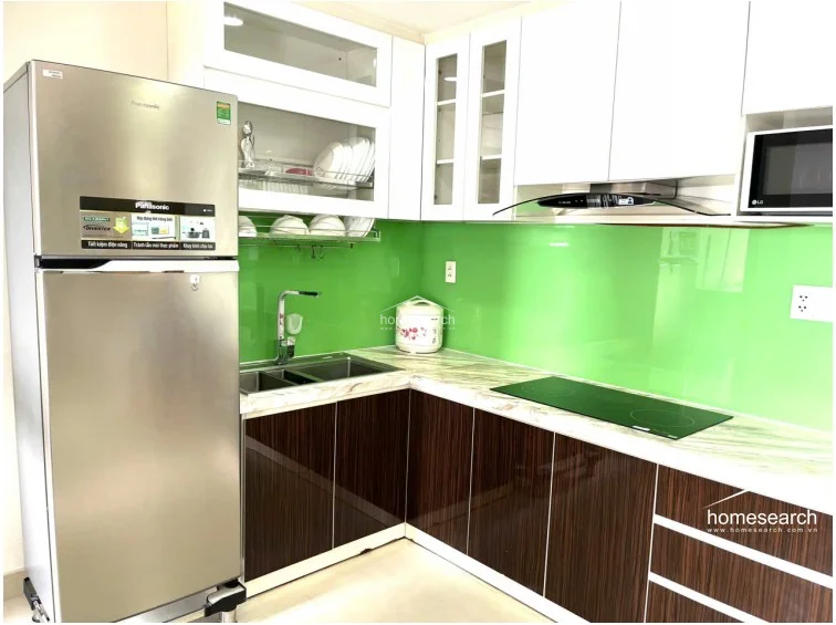 This L-shaped kitchen features a modern aesthetic with high-contrast dark wood-grain lower cabinets and crisp white upper cabinetry. The space is highlighted by a vibrant green glass backsplash and a marble-patterned countertop, equipped with a stainless steel refrigerator and an integrated induction cooktop.