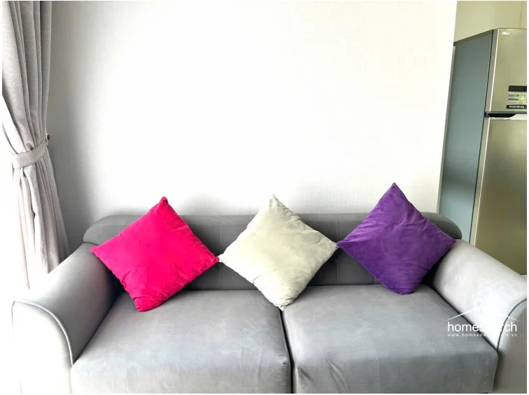 This living area features a comfortable grey sofa accented with vibrant pink, cream, and purple throw pillows, set against a neutral wall. A stainless steel refrigerator is visible in the adjacent kitchen area, suggesting an open-concept flow with a contemporary, minimalist aesthetic.
