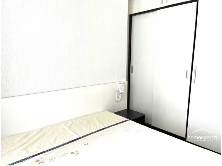 This bedroom features a minimalist design with a built-in white sliding-door wardrobe that maximizes floor space. The room includes a wall-mounted bedside lamp and neutral, textured wall finishes, creating a clean and organized sleeping environment.