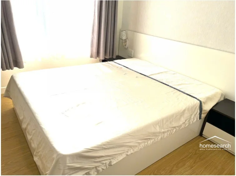 This bedroom is furnished with a large bed featuring a white headboard and neutral bedding, complemented by a dark-toned nightstand. The room includes light wood-look flooring and floor-to-ceiling curtains that allow for soft natural light, creating a simple and well-maintained sleeping environment.