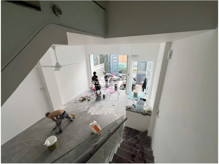 This view from the staircase captures the open-plan ground floor, which flows directly toward the gated entrance and outdoor area. The space features large-format grey floor tiles, crisp white walls, and a modern ceiling fan, currently undergoing final cleaning and finishing work.