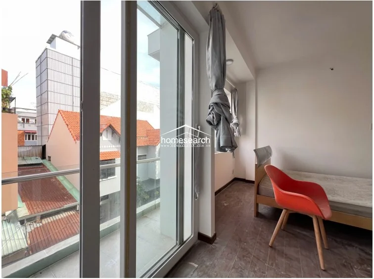 This bedroom view highlights the connection to a private balcony through large sliding glass doors, which allow for ample natural light. The room is finished with dark wood-toned flooring and features a simple, modern aesthetic with a red chair providing a pop of color.