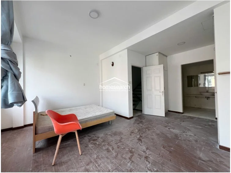 This bedroom features dark wood-look flooring and a simple, minimalist layout with a single bed and a vibrant red accent chair. The room includes a private ensuite bathroom and a built-in wardrobe, maintaining a neutral white color scheme throughout.