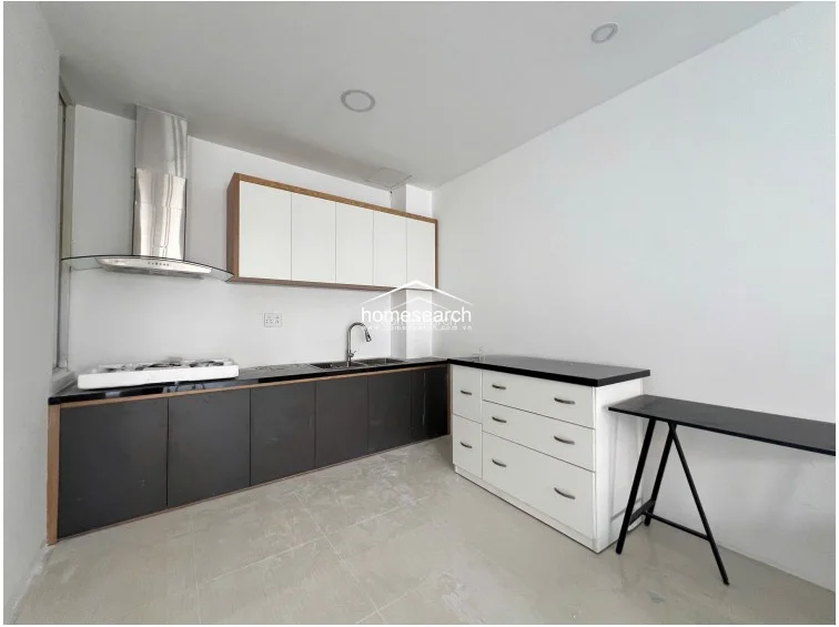 This minimalist kitchen features a linear layout with dark lower cabinets, white upper cabinetry, and a black countertop. It is equipped with a modern stainless steel range hood and a gas stovetop, set against a clean white wall finish and light-colored tiled flooring.