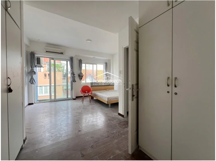 This spacious bedroom features a neutral color palette with white walls and dark wood-look flooring, creating a clean, minimalist aesthetic. It includes a private balcony with glass railings, a wall-mounted AC unit, and extensive built-in white wardrobes for ample storage.