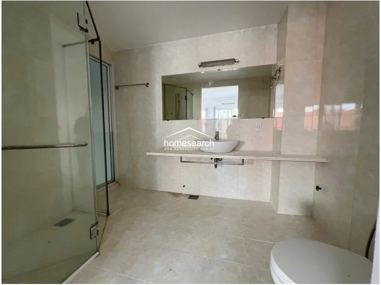 This spacious, minimalist bathroom features a wide vanity counter with a vessel sink and a large, full-width wall mirror. It includes a glass-enclosed shower area and is finished with light-toned wall and floor tiles, creating an airy and well-lit atmosphere.
