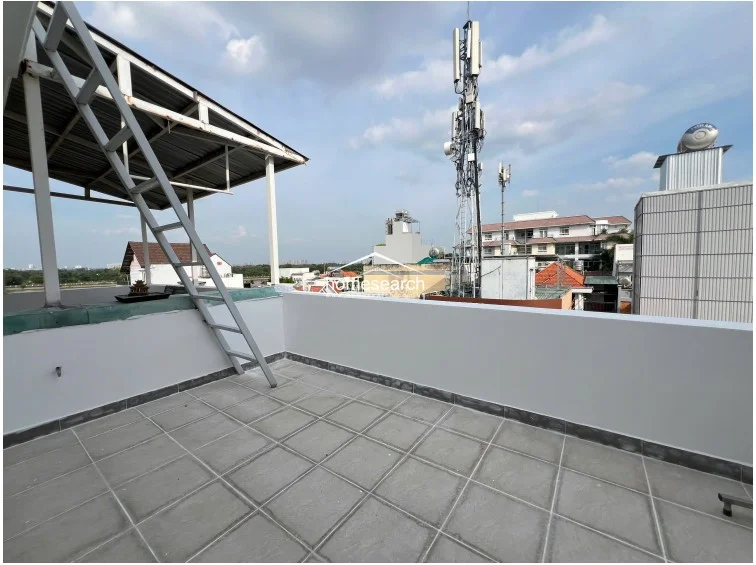 This open-air rooftop terrace provides a functional outdoor space with light-colored floor tiles and a partial metal roof structure. It offers an urban view of the surrounding neighborhood, including nearby buildings and telecommunication infrastructure.