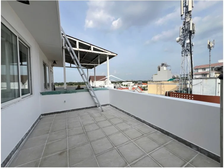 This spacious rooftop terrace is paved with large grey tiles and features a white perimeter wall, offering an open-air setting with expansive city views. A metal ladder structure provides access to an elevated roof section, and the area is well-maintained, providing a versatile outdoor space for relaxation or utility.