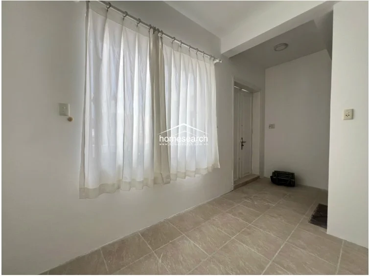 This room is finished with light-colored floor tiles and neutral white walls, creating a clean and bright environment. It features a large window dressed with white curtains and leads into a secondary space through a doorway, suggesting a functional, multi-purpose layout.
