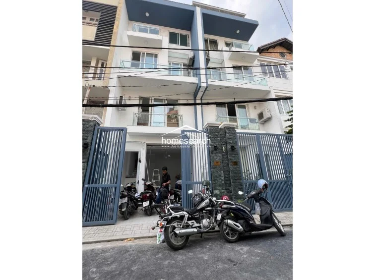 This modern multi-story townhouse features a contemporary facade with clean lines, blue and white accents, and glass-railed balconies on each level. The property includes a secure gated entrance with stone detailing and appears to be in excellent, newly constructed condition.