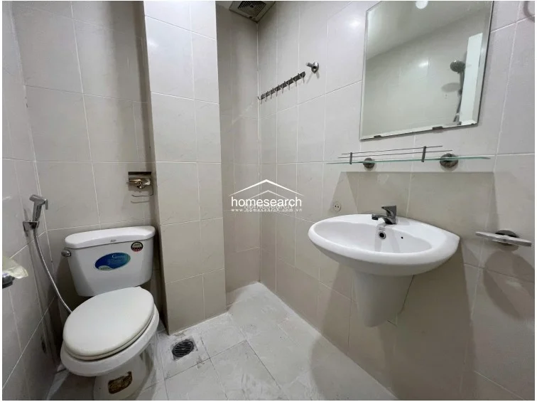 This compact bathroom features floor-to-ceiling light-colored tiling and a pedestal sink with a wall-mounted mirror. It includes a standard toilet, a handheld bidet sprayer, and a wall-mounted rack for storage, maintaining a clean and functional condition.