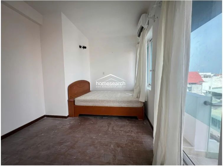 This bedroom offers a bright, airy atmosphere with large windows that provide natural light and a view of the neighborhood. It is furnished with a wooden bed frame, a wall-mounted AC unit, and a modern black wall sconce, all set against a backdrop of white walls and dark flooring.