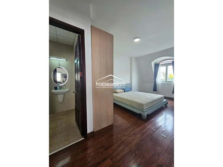 This bedroom features a unique architectural archway window and rich, dark wood-tone flooring that adds warmth to the space. It includes an en-suite bathroom with a modern, illuminated circular mirror and a wall-mounted sink, reflecting a well-maintained and contemporary interior.