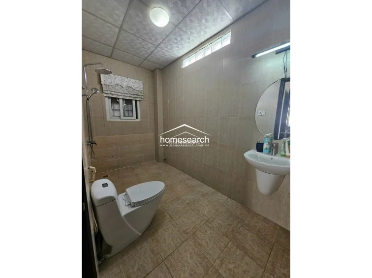 This bathroom is finished with neutral-toned wall and floor tiles and features a pedestal sink paired with a large circular mirror. A small high-level window with a patterned blind provides natural light, while the ceiling is finished with a drop-panel design.