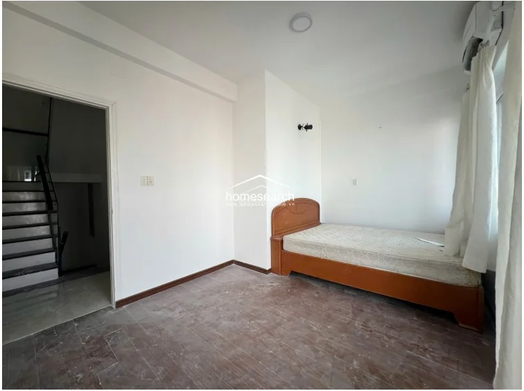 This bedroom features a simple layout with white walls and dark wood-look flooring that appears to be in need of a deep clean. It includes a basic wooden bed frame and is equipped with a wall-mounted AC unit, while the doorway provides direct access to an adjacent stairwell.