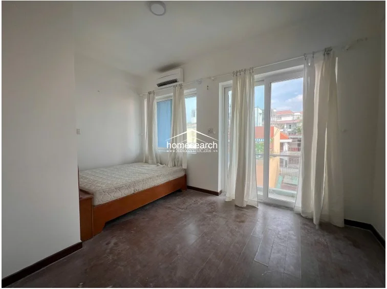 This bedroom space is brightened by large glass doors that offer access to an outdoor area and allow for significant natural light. The room is sparsely furnished with a single bed and an AC unit, maintaining a minimalist aesthetic with white walls and dark flooring.