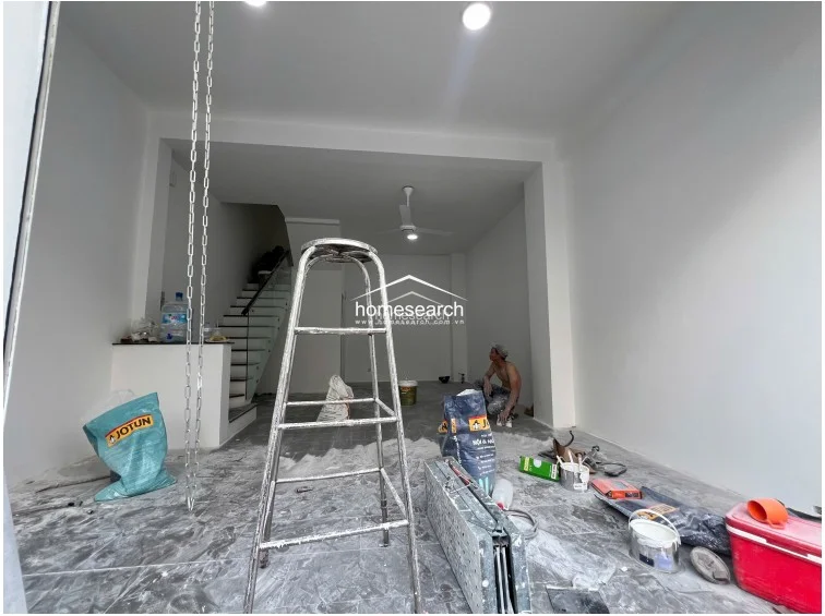 This ground-floor living area shows a minimalist, open layout that connects seamlessly to the staircase and the front entrance. The space is illuminated by recessed ceiling lights and a ceiling fan, with neutral white walls and grey tiled flooring providing a clean, modern aesthetic.