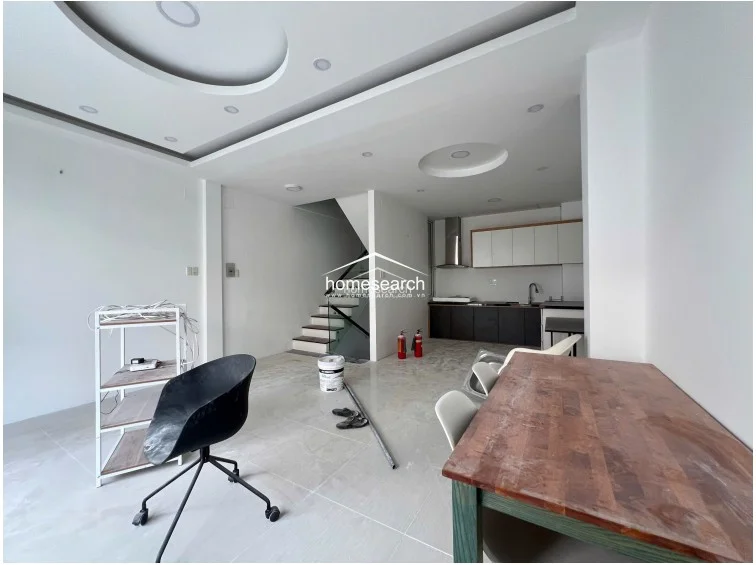 This open-concept living and kitchen area features bright white walls and polished light-colored tile flooring, creating a clean and modern atmosphere. The space is anchored by a contemporary kitchen with two-tone cabinetry and a stainless steel range hood, while a staircase with a sleek glass railing provides a direct flow to other levels.