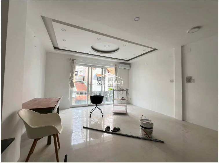 This view highlights the living area's connection to a private balcony through large glass sliding doors, which allow for ample natural light and ventilation. The room is equipped with a wall-mounted AC unit and features a sophisticated recessed ceiling design with integrated lighting, emphasizing the property's well-maintained, minimalist aesthetic.