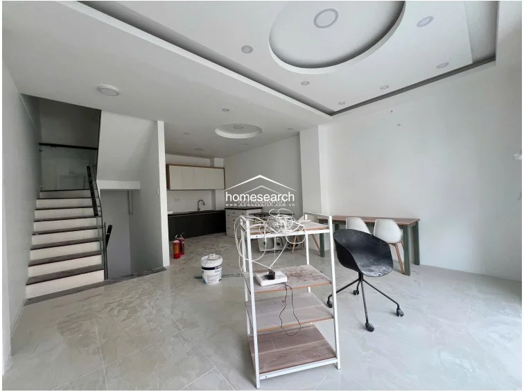 This perspective shows the transition between the living space and the kitchen, highlighting the functional layout and the staircase as a central architectural feature. The neutral color palette and consistent tile flooring throughout unify the space, while the presence of fire extinguishers indicates attention to safety.
