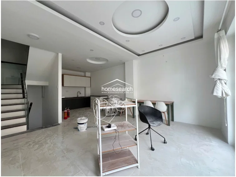 This open-plan living and dining area features a modern kitchen at the rear with dark lower cabinetry and white upper cupboards. The space is highlighted by a sophisticated recessed ceiling with circular lighting features, polished light-colored floor tiles, and an open staircase with a glass railing.