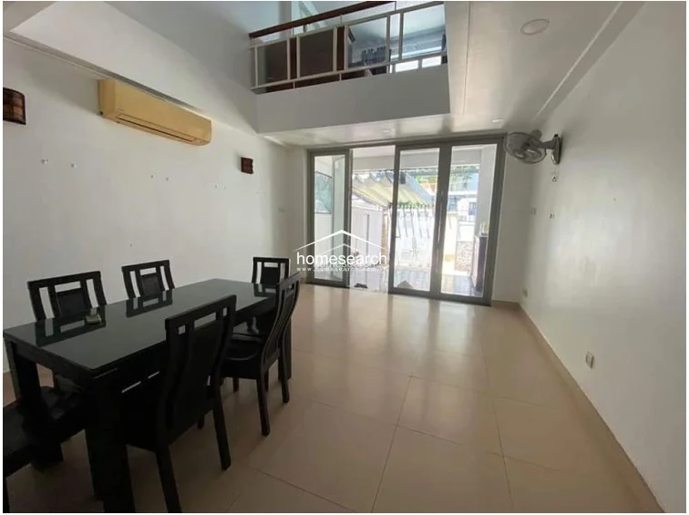 This dining area features a minimalist, open-plan layout with light-colored tile flooring and white walls, creating a bright and airy atmosphere. The space is equipped with a wall-mounted AC unit and a ceiling fan, and it connects directly to the outdoor courtyard through large glass sliding doors, allowing for excellent natural light.
