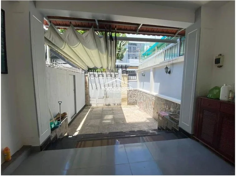 This outdoor courtyard serves as a functional entrance or parking area, featuring stone-tiled flooring and a retractable fabric awning for shade. The space is enclosed by high walls with stone cladding accents and includes a secure metal gate, reflecting a well-maintained, private exterior area.