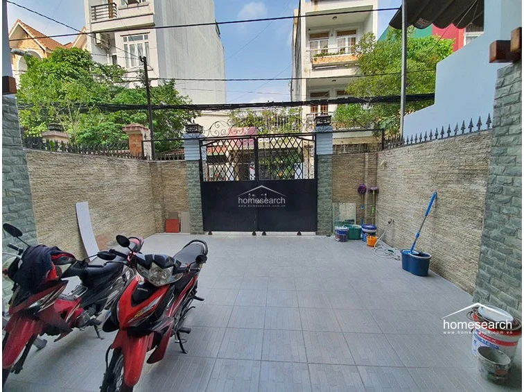 This outdoor courtyard serves as a secure, gated parking area for motorbikes, featuring durable light-grey floor tiles and textured stone wall cladding. The space is enclosed by a tall metal gate and high perimeter walls, providing a functional and private transition from the street.