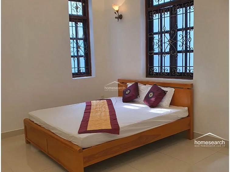 This bedroom offers a simple, functional layout centered around a wooden bed frame positioned between two windows with decorative metal grilles. The room is illuminated by a warm wall-mounted sconce and maintains a bright, airy feel with its neutral wall finish and polished tile floors.