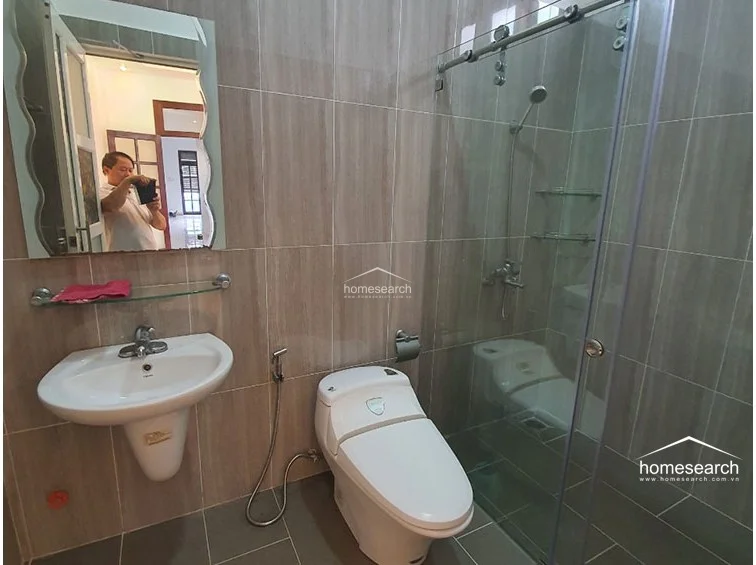 This bathroom features a contemporary design with neutral, wood-look wall tiling and a glass-enclosed shower stall. It is equipped with a modern pedestal sink, a wall-mounted mirror with decorative edges, and a sleek, dual-flush toilet.