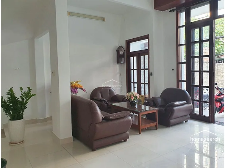 This living area features a clean, minimalist layout with white walls and polished light-colored tile flooring, creating a bright and airy atmosphere. The space is furnished with a dark leather sofa set and a glass-top coffee table, connecting directly to the exterior through a large, multi-paned glass door that allows for ample natural light.