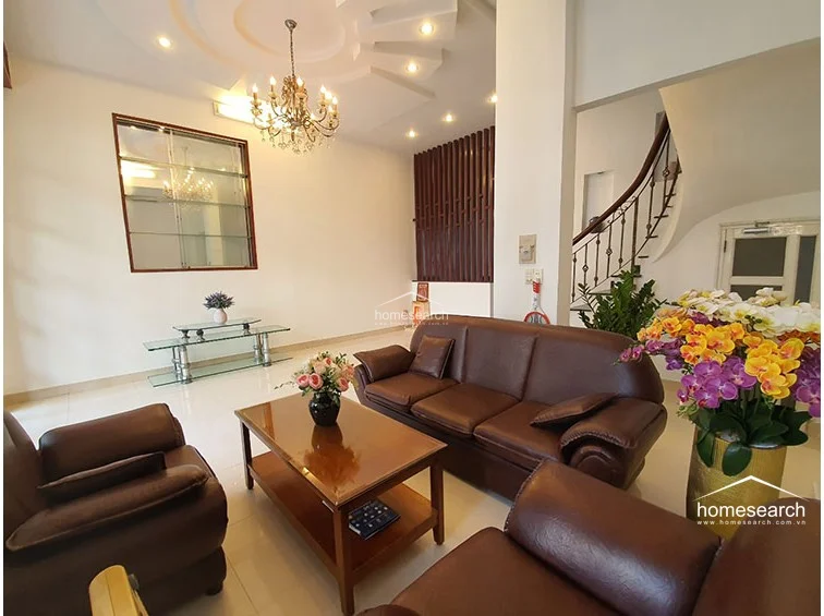 This spacious living room is defined by a sophisticated tray ceiling with recessed lighting and an elegant central chandelier. The room features a classic dark leather seating arrangement and a decorative glass display shelf, with a sweeping staircase in the background that adds architectural interest and flow to the upper levels.