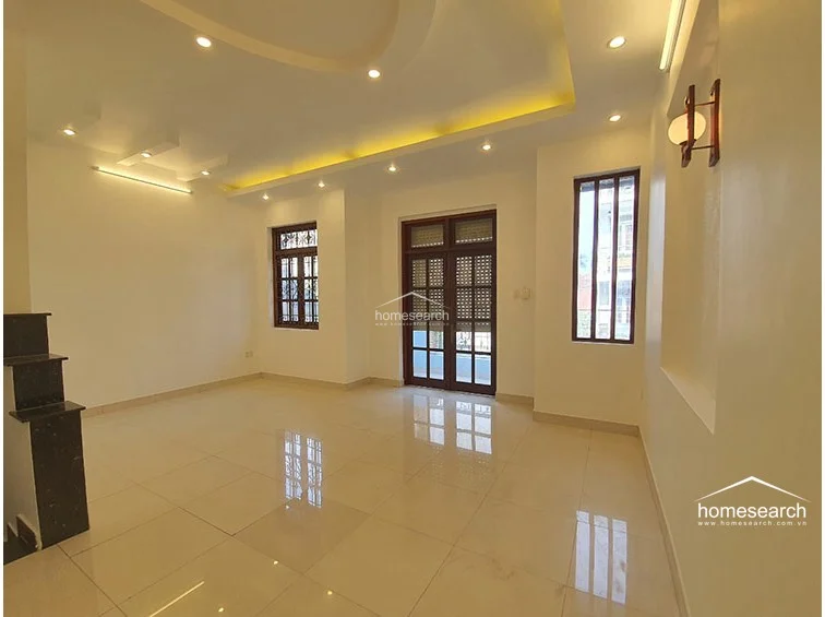 This spacious, open-plan living area features high ceilings with recessed cove lighting and a neutral color palette that enhances the sense of space. The room is finished with polished light-colored floor tiles and includes a central doorway leading to an outdoor balcony, flanked by two tall windows with dark wood frames and decorative security grilles.
