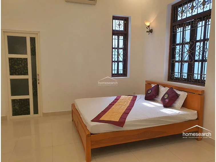 This bedroom features a light wood bed frame dressed with a traditional runner, set against plain white walls and polished tile flooring. The room is equipped with a wall-mounted air conditioning unit and receives natural light through a window adorned with intricate metal security bars.