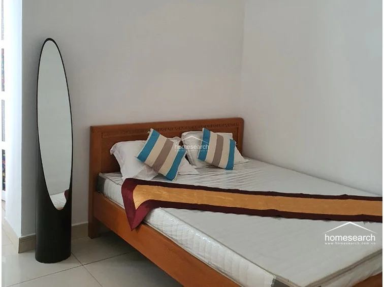 This bedroom layout highlights a minimalist design with a wooden bed frame and a full-length, oval-shaped floor mirror positioned in the corner. The space maintains a clean, well-maintained aesthetic with white walls and consistent light-colored floor tiles throughout.