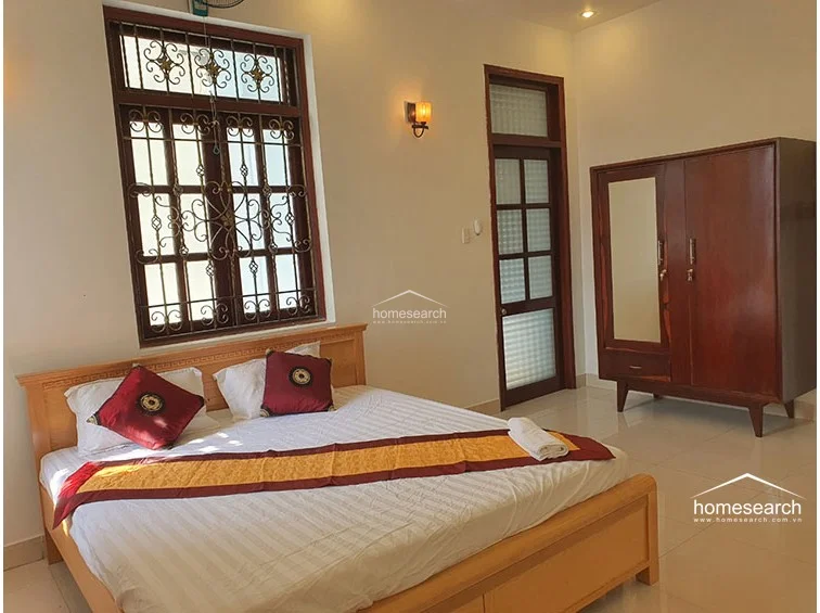 This bedroom is furnished with a wooden bed and a matching freestanding wardrobe, providing essential storage in a clean, uncluttered setting. The room features a frosted glass door for privacy and a large window with ornate security ironwork, ensuring both ventilation and natural light.