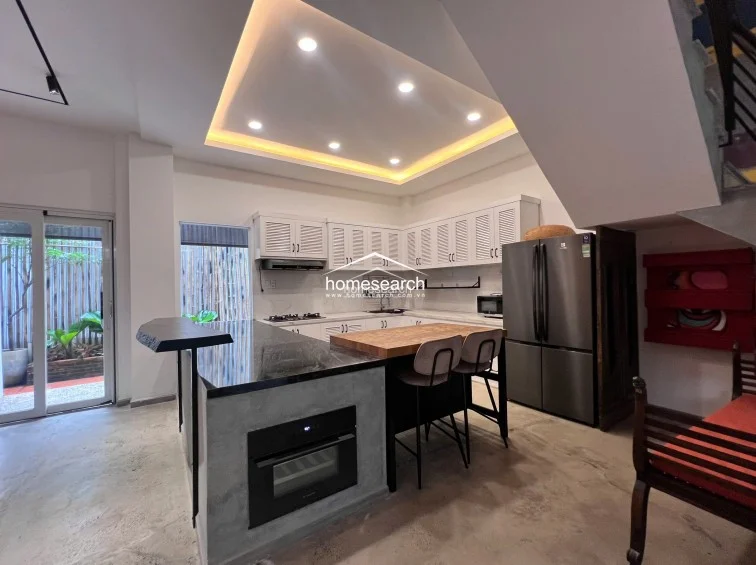 This open-concept kitchen features a U-shaped layout with white louvered cabinetry, marble-look countertops, and a sleek black stone island equipped with a built-in oven. The space is well-lit by recessed ceiling lights and a cove-lit tray ceiling, while a large stainless steel refrigerator and modern stove complete the high-end appliance suite.