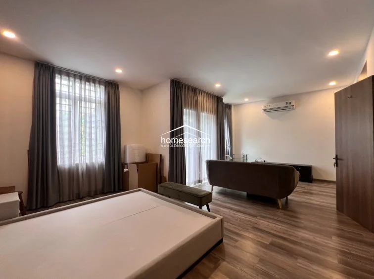 This bedroom layout emphasizes a clean, minimalist aesthetic with wood-look flooring and a wall-mounted air conditioning unit for climate control. The room is bright and airy, featuring large windows dressed in floor-to-ceiling curtains and a comfortable seating area, suggesting a well-maintained and relaxing environment.
