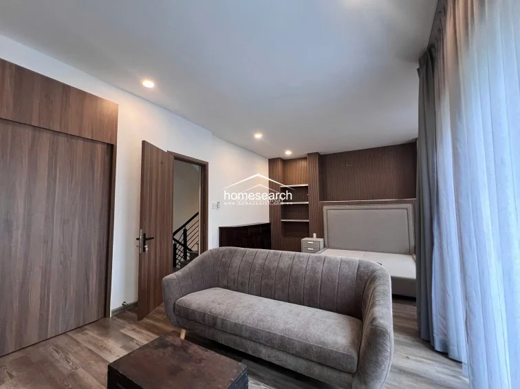 This spacious bedroom features a modern, neutral color palette with warm wood-look flooring and matching wood-veneer built-in cabinetry. The room is furnished with a comfortable grey sofa and a plush upholstered bed, while recessed ceiling lighting provides a clean, contemporary aesthetic. A doorway leads directly to a staircase landing, suggesting a convenient and accessible layout.