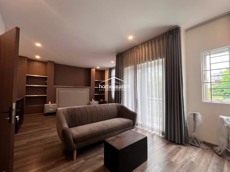 This bedroom layout emphasizes comfort and storage, featuring a large bed with a matching upholstered headboard and integrated shelving units on either side. The room is brightened by floor-to-ceiling curtains that cover a balcony door, allowing for ample natural light and outdoor access. The space is well-maintained, with a cohesive design that pairs dark wood textures against soft, neutral walls.