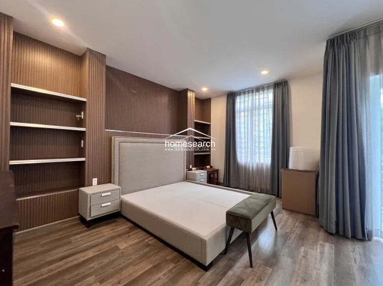 This well-appointed bedroom includes a queen-sized bed with a fabric headboard and a stylish, low-profile green bench at the foot. The room is framed by floor-to-ceiling curtains and built-in wooden shelving, creating a sophisticated and organized atmosphere. The warm wood flooring and recessed lighting contribute to a cozy, high-end feel throughout the space.