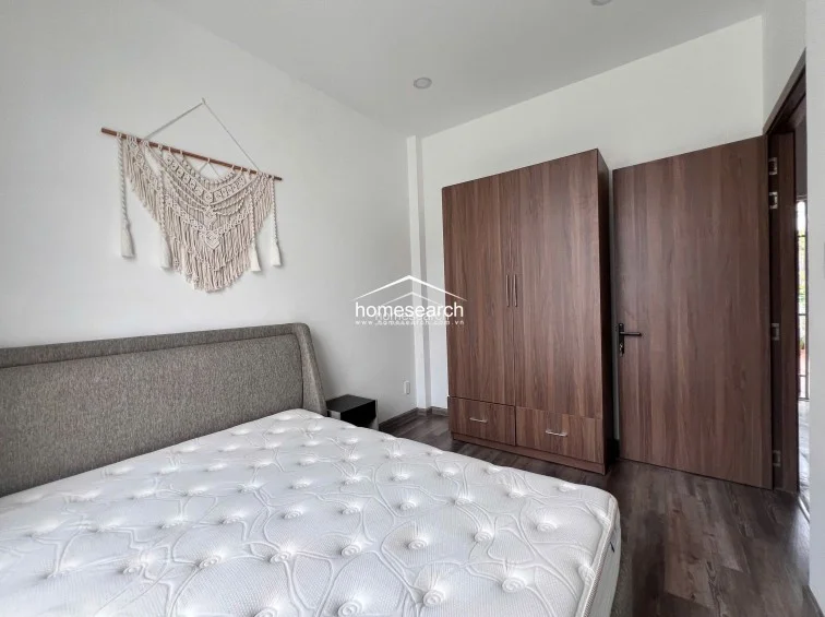 This compact bedroom is designed with efficiency in mind, featuring a queen-sized bed and a tall, wood-finish wardrobe that offers substantial vertical storage. The minimalist white walls are accented by a decorative macramé wall hanging, adding a touch of texture to the clean, modern aesthetic. The room is finished with dark wood-look flooring and a matching wooden door, maintaining a consistent and polished design theme.