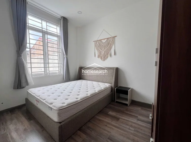 This cozy bedroom features a neutral color palette with white walls and dark wood-look flooring, creating a clean and modern aesthetic. The room is anchored by a grey upholstered bed frame and a decorative macramé wall hanging, while a large window with security bars provides natural light and grey curtains for privacy.