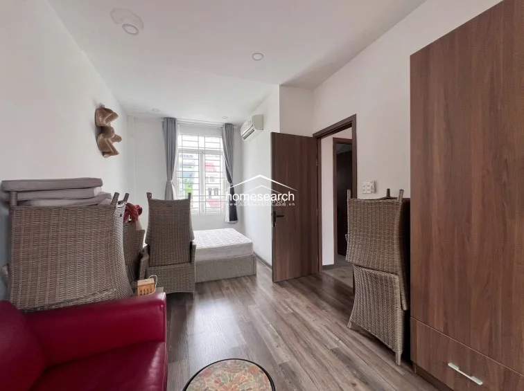 This bedroom space is currently being used for storage, with several wicker chairs stacked and a red sofa positioned near the entrance. The room includes a tall wooden wardrobe for storage, a wall-mounted air conditioning unit, and a large window that allows for ample natural light.