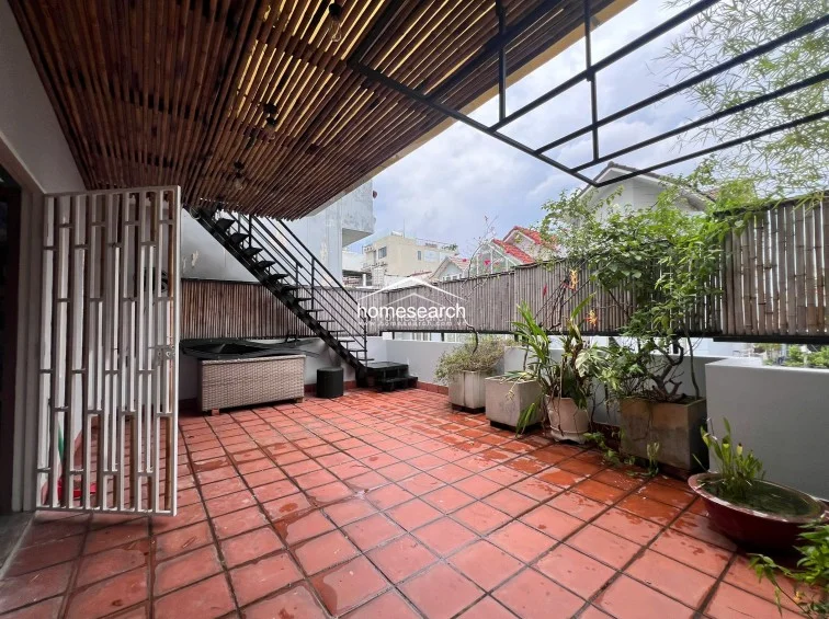 This spacious outdoor terrace is paved with terracotta-style tiles and features a bamboo-slatted pergola that provides partial shade. The area is well-maintained with various potted plants and includes a metal staircase leading to an upper level, offering a functional and relaxing outdoor retreat.