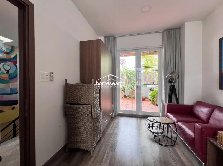 This bright living area connects directly to the outdoor terrace through double glass doors, promoting a seamless indoor-outdoor flow. The room is furnished with a vibrant red sofa, a tall wooden wardrobe for storage, and a modern floor lamp, all set against a backdrop of light walls and dark wood-look flooring.